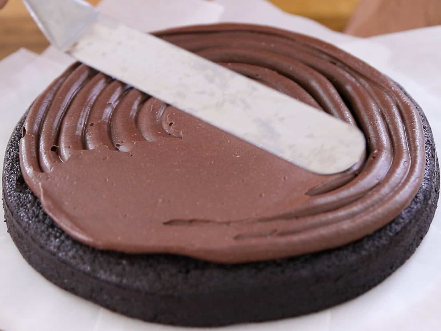 A close-up of a spatula spreading chocolate frosting onto a round, dark chocolate cake layer on parchment paper.