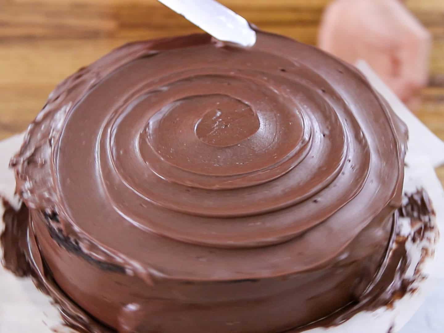 A hand uses a spatula to smooth chocolate frosting on a round cake, creating concentric circular patterns on the top.