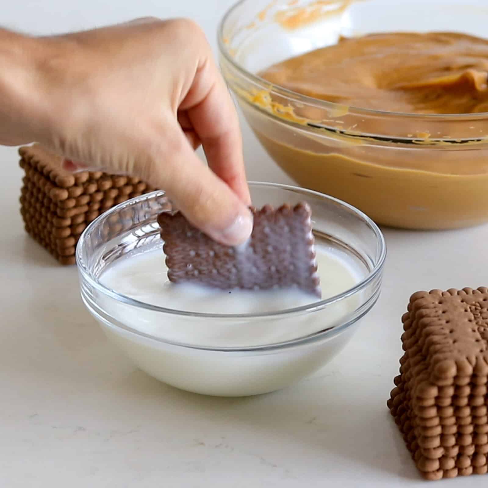 A hand dips a rectangular chocolate biscuit into a glass bowl of milk. Stacks of biscuits and a bowl of caramel-colored mixture are on the counter in the background.
