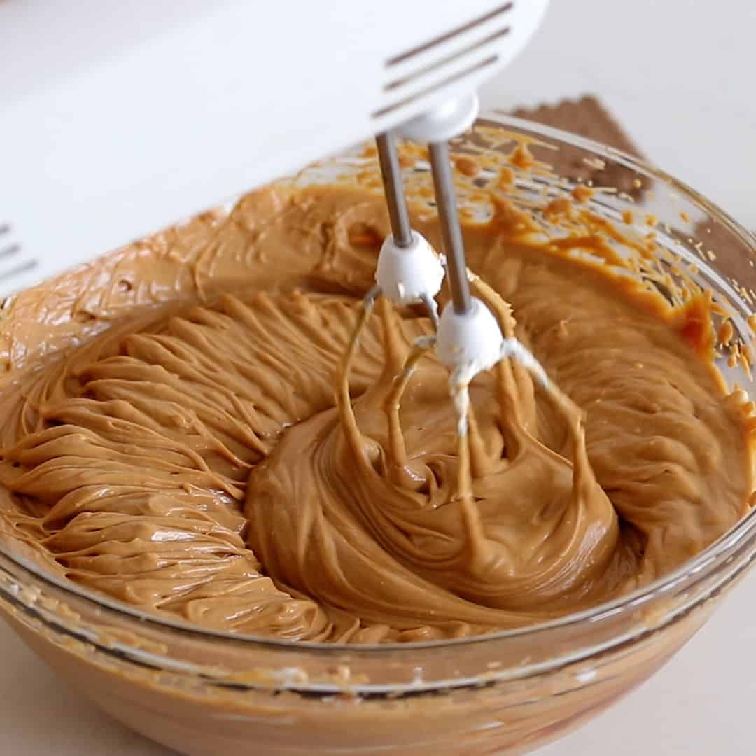 A close-up of an electric hand mixer blending creamy, light brown batter in a glass bowl on a kitchen countertop.
