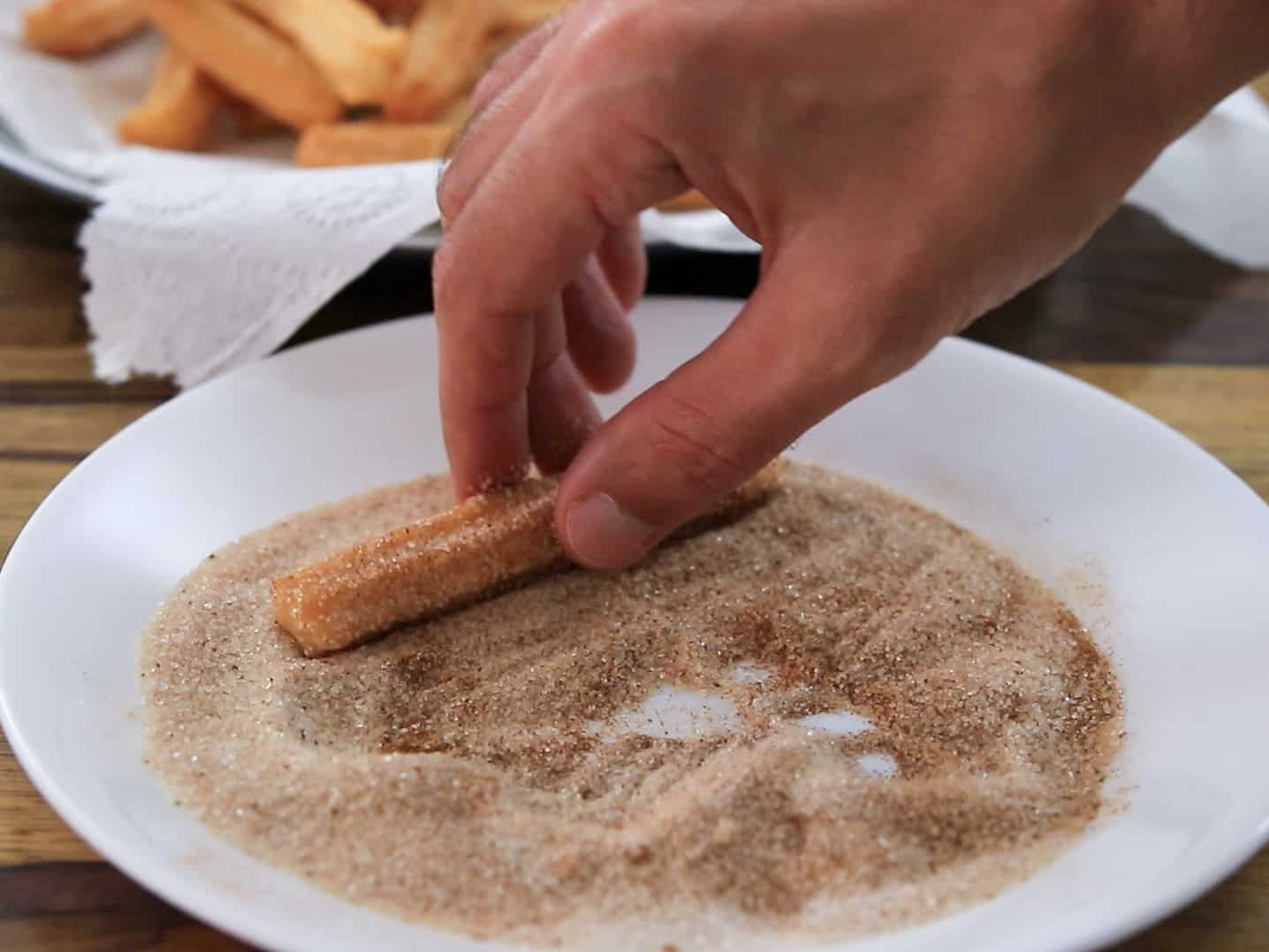A hand rolls a churro stick in a plate of cinnamon sugar, with more churros on a napkin-covered plate in the background.