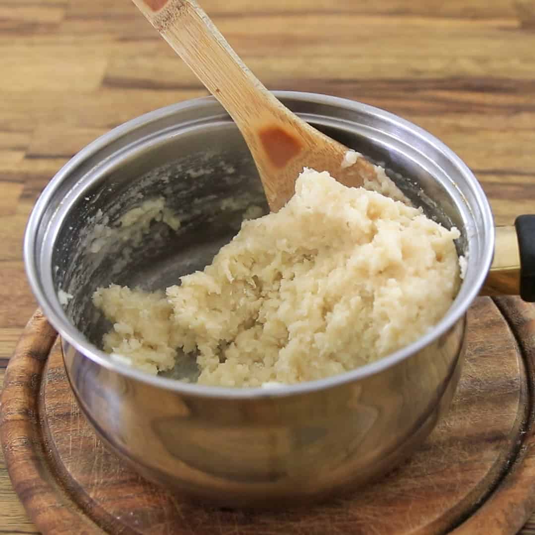 A stainless steel pot containing churros dough on a wooden cutting board, with a wooden spoon resting inside the pot.