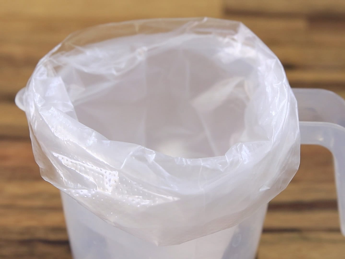 A transparent plastic bag is spread open and fitted over the rim of a clear plastic pitcher, with a wooden surface in the background.