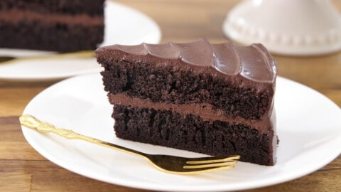 A slice of rich, layered chocolate cake with chocolate frosting sits on a white plate with a gold fork beside it, set on a wooden table.