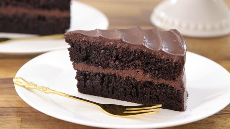 A slice of rich, layered chocolate cake with chocolate frosting sits on a white plate with a gold fork beside it, set on a wooden table.