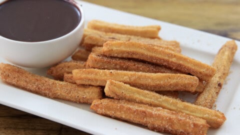 A plate of golden-brown churros coated with sugar, served next to a small bowl of thick chocolate sauce for dipping.