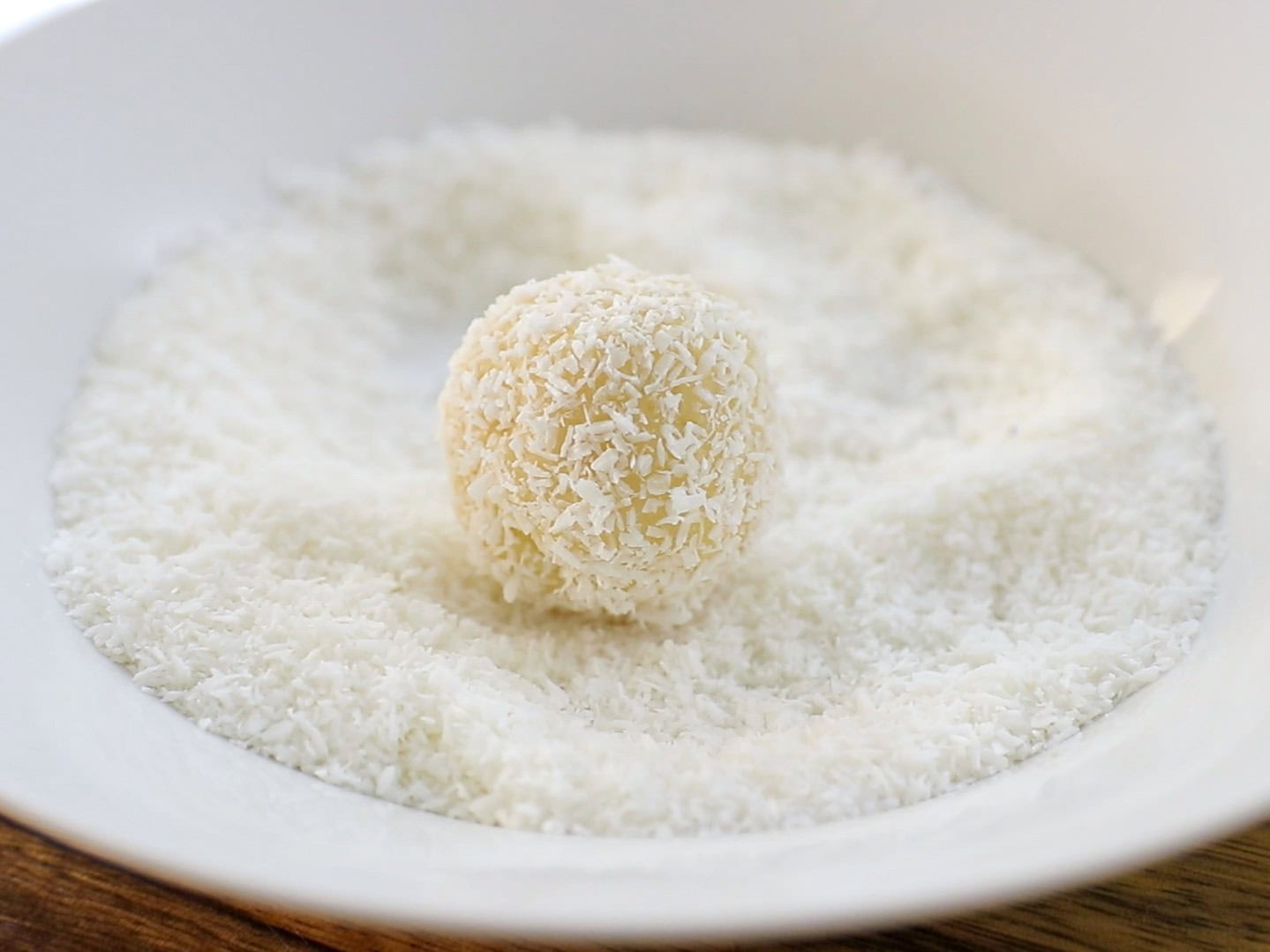 A single round white coconut white chocolate truffles sits in a white bowl filled with shredded coconut, with a wooden surface partially visible underneath.