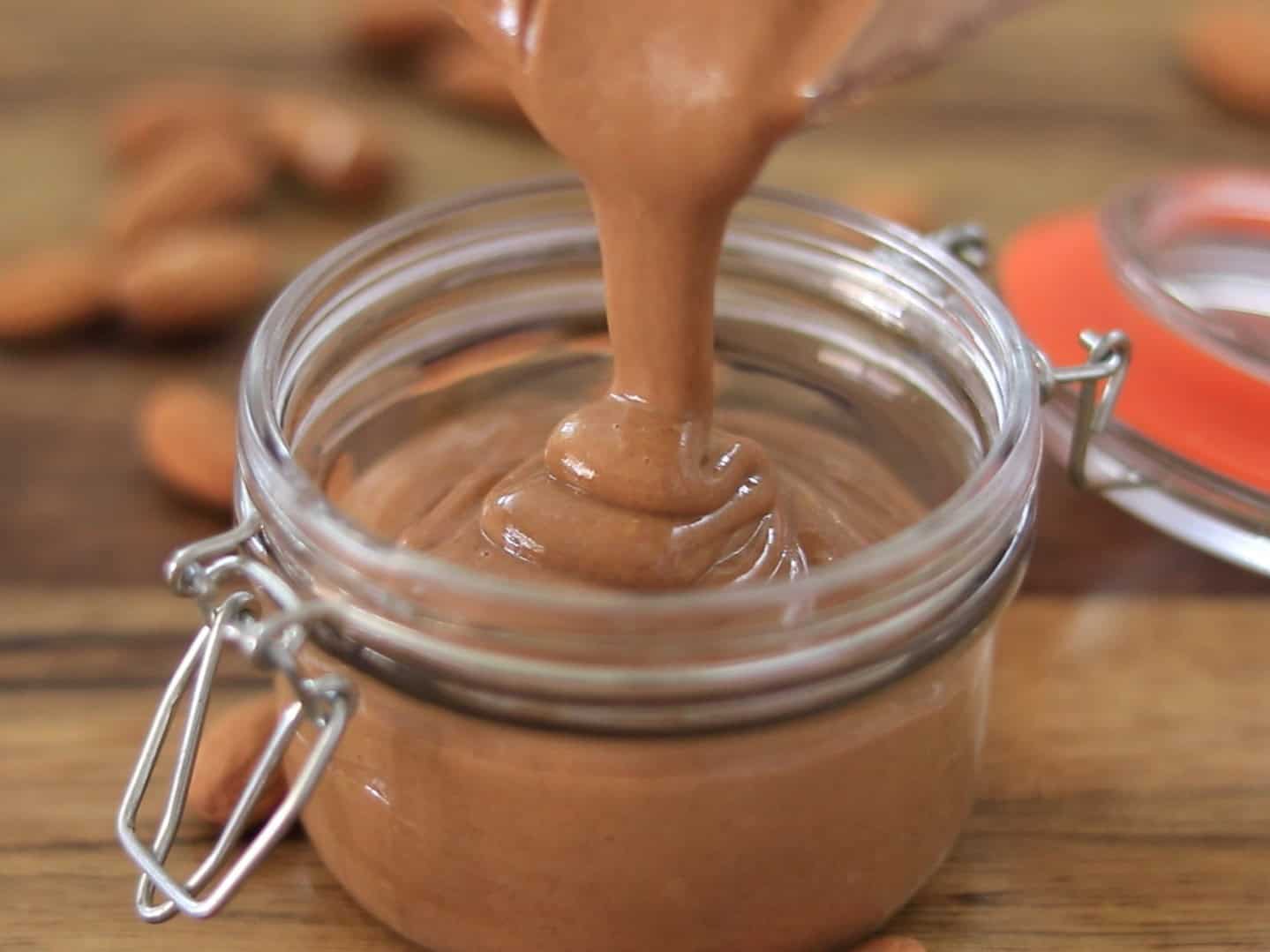A thick, creamy almond butter is being poured into an open glass jar with a metal clasp, surrounded by scattered whole almonds on a wooden surface.
