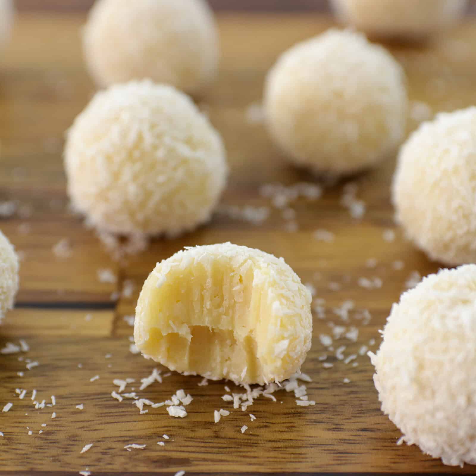 Several round white chocolate coconut truffles are on a wooden surface, with one truffle in the foreground showing a bite taken out, revealing the creamy interior. Shredded coconut is sprinkled around them.