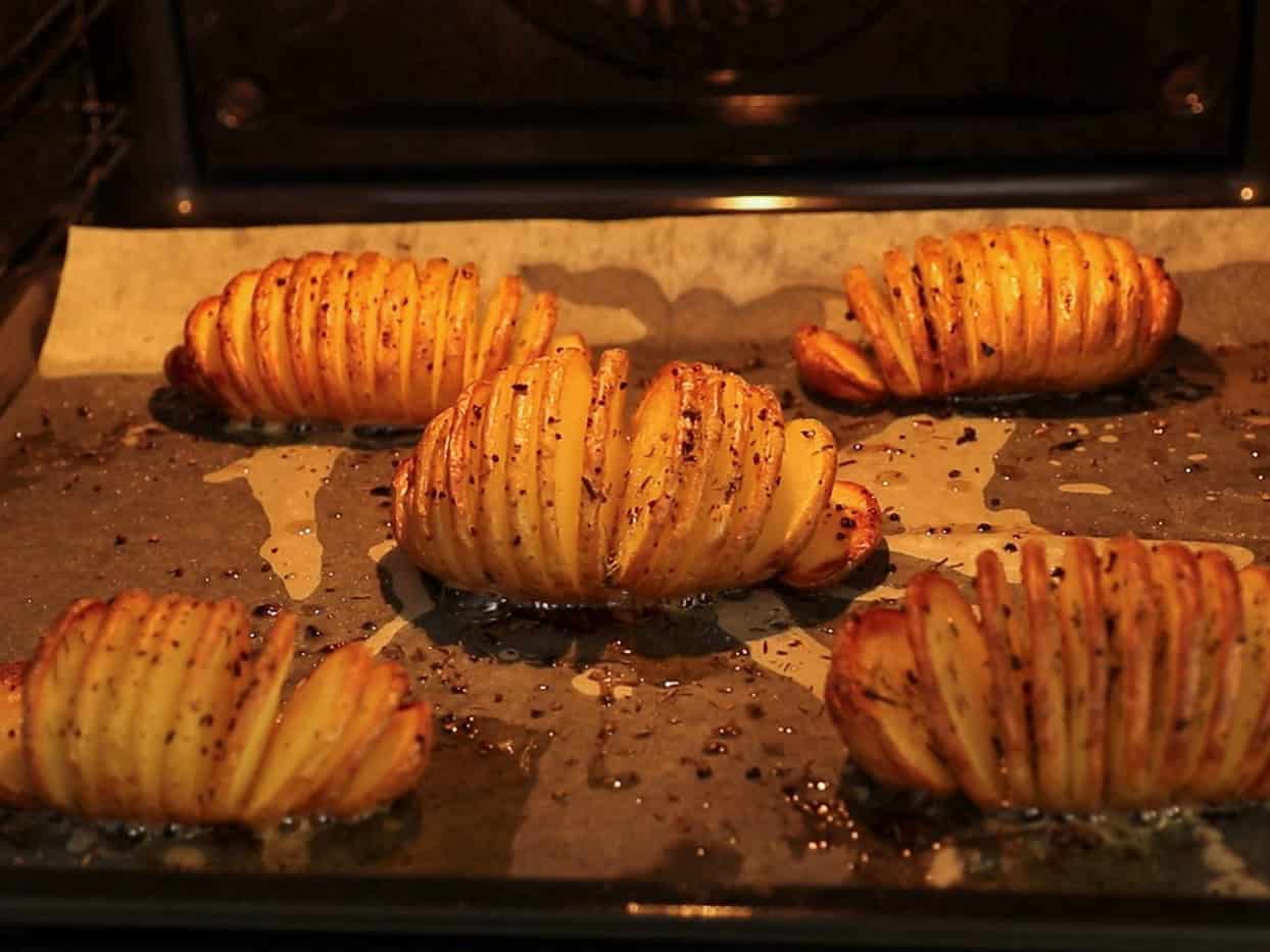 Five sliced, seasoned Hasselback potatoes are baking on a parchment-lined tray in an oven, with crispy edges and oil glistening on the surface.