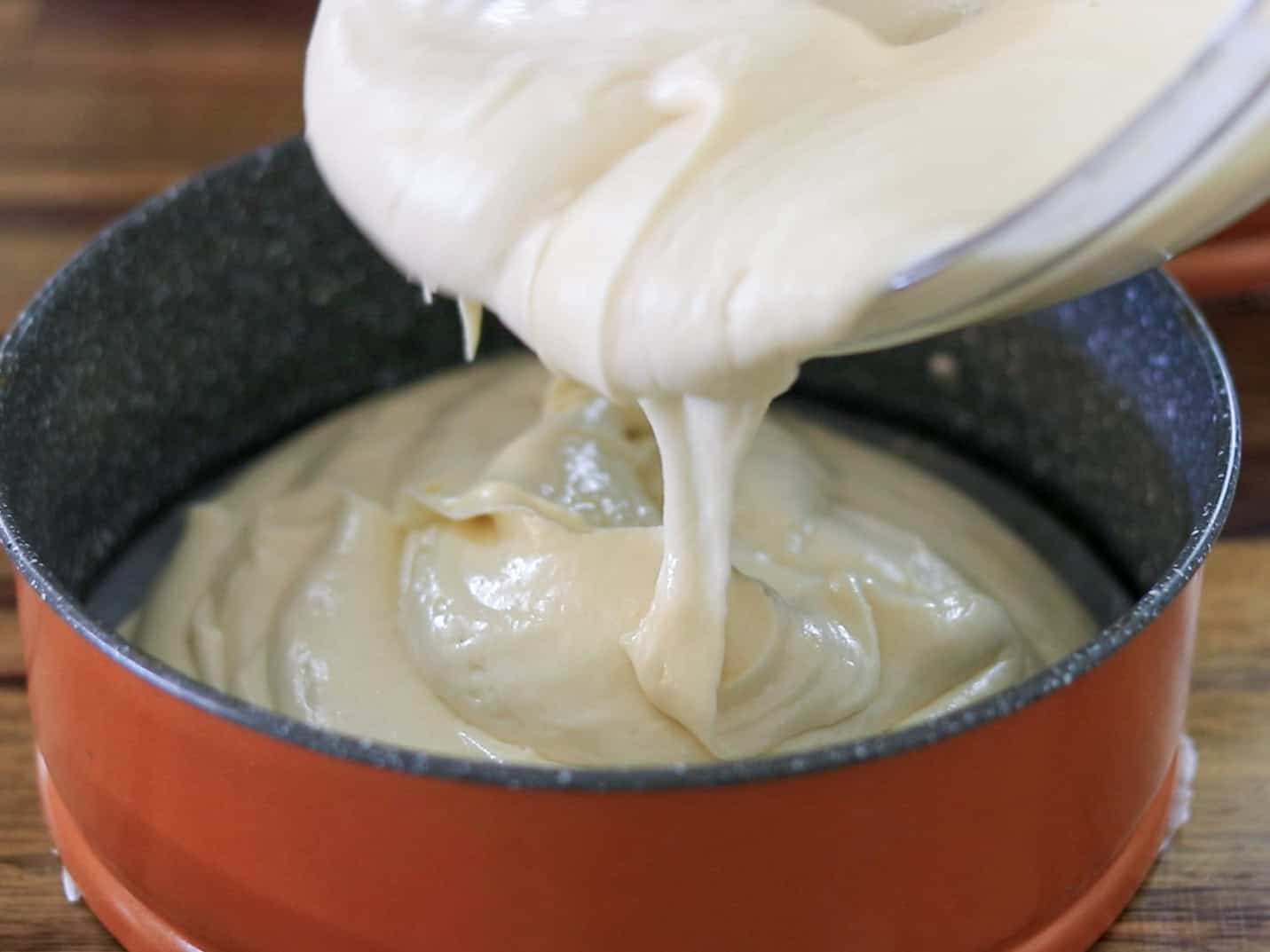Thick cake batter is being poured from a bowl into a round, orange baking pan lined with parchment paper, ready for baking.