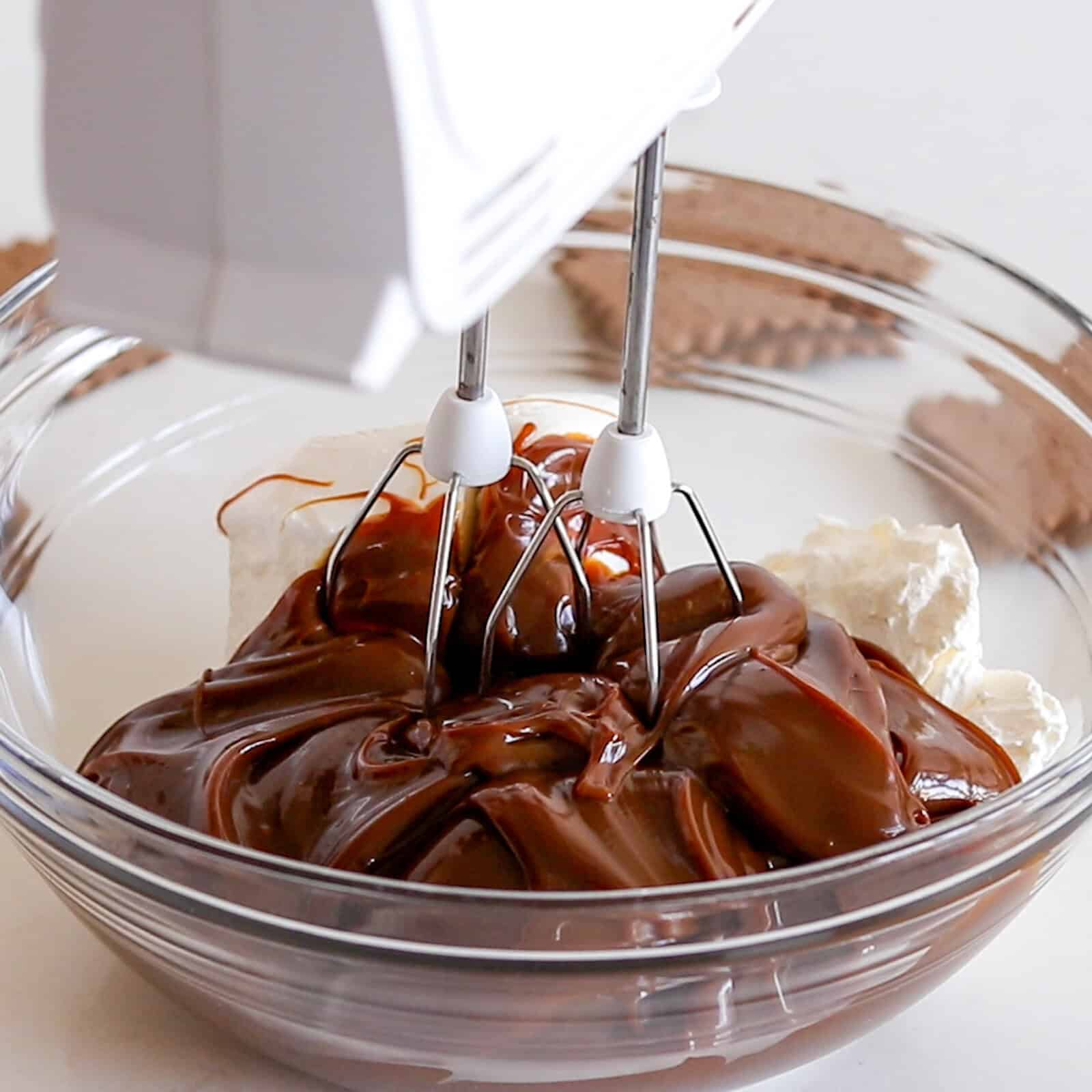 A hand mixer with beaters is mixing chocolate and cream cheese in a clear glass bowl. Chocolate cookies are visible in the background on a white surface.