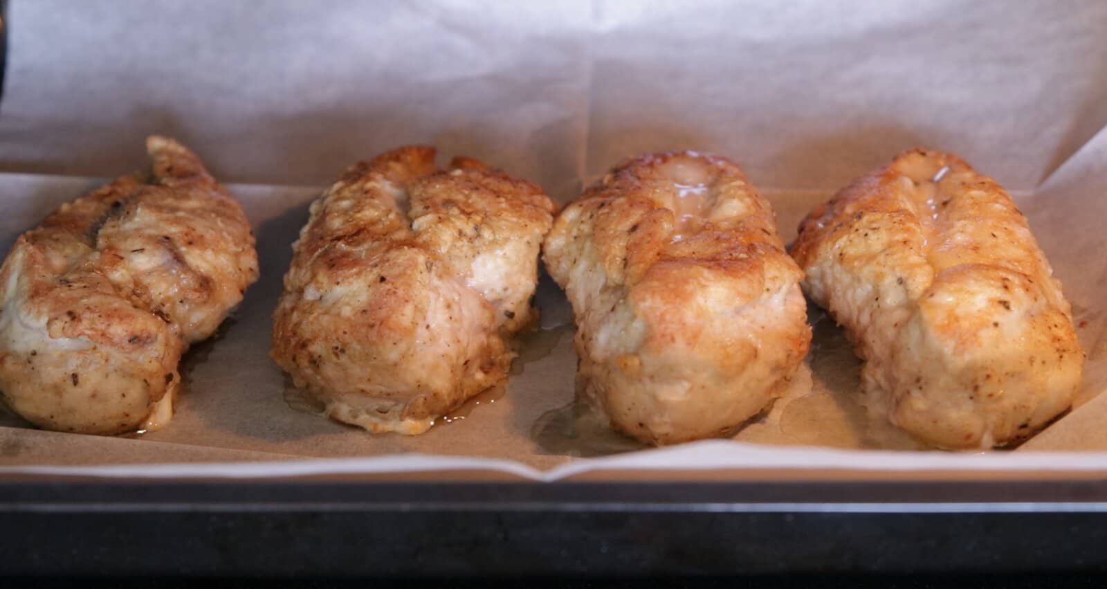 Four browned, cooked chicken breasts on a baking tray lined with parchment paper, fresh from the oven.