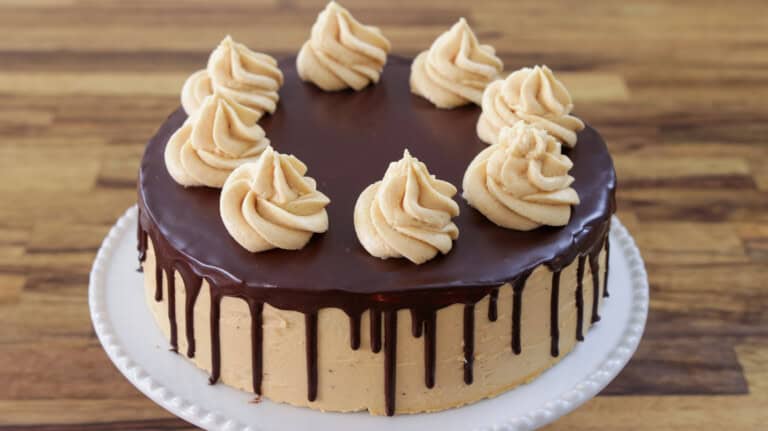 A round cake with light brown frosting and chocolate ganache dripping down the sides, topped with eight swirls of frosting, displayed on a white plate with a wooden background.