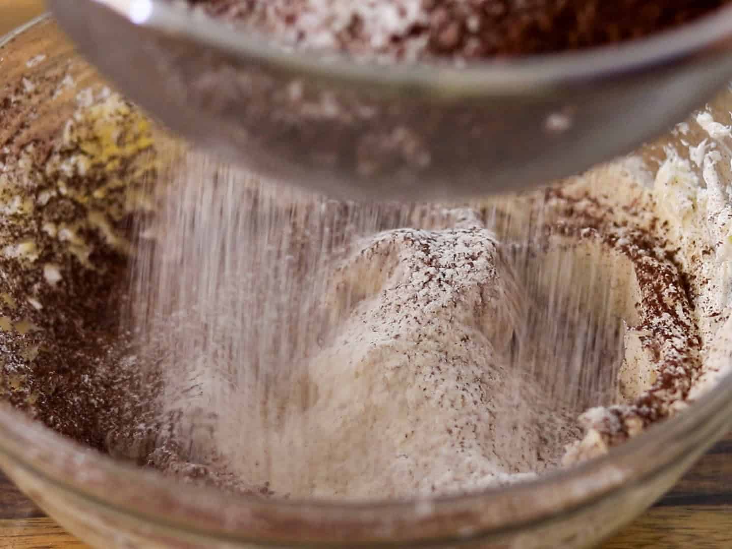 Cocoa powder is being sifted from a bowl onto a mound of flour in a glass mixing bowl, ready for baking.