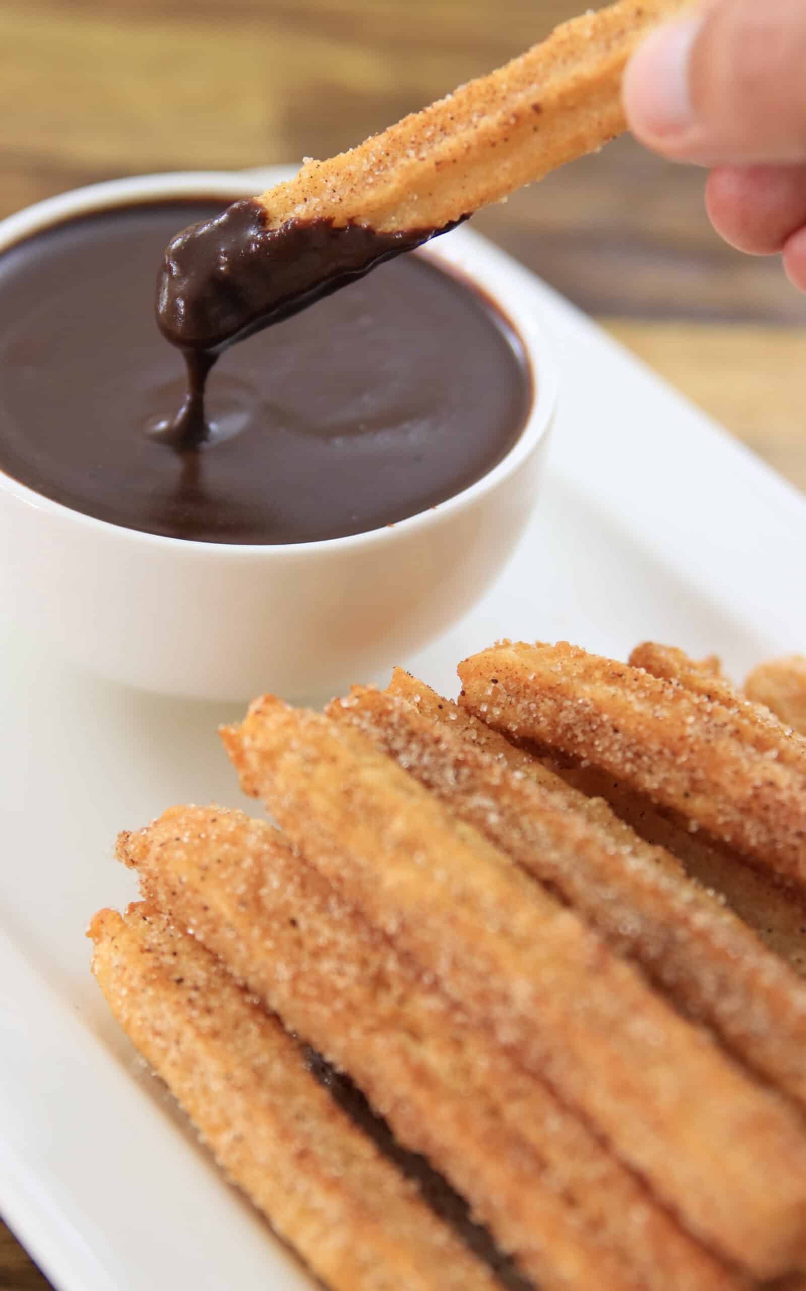 A hand dips a cinnamon sugar-coated churro into a bowl of thick, dark chocolate sauce. Several churros are stacked on a white plate in the foreground.