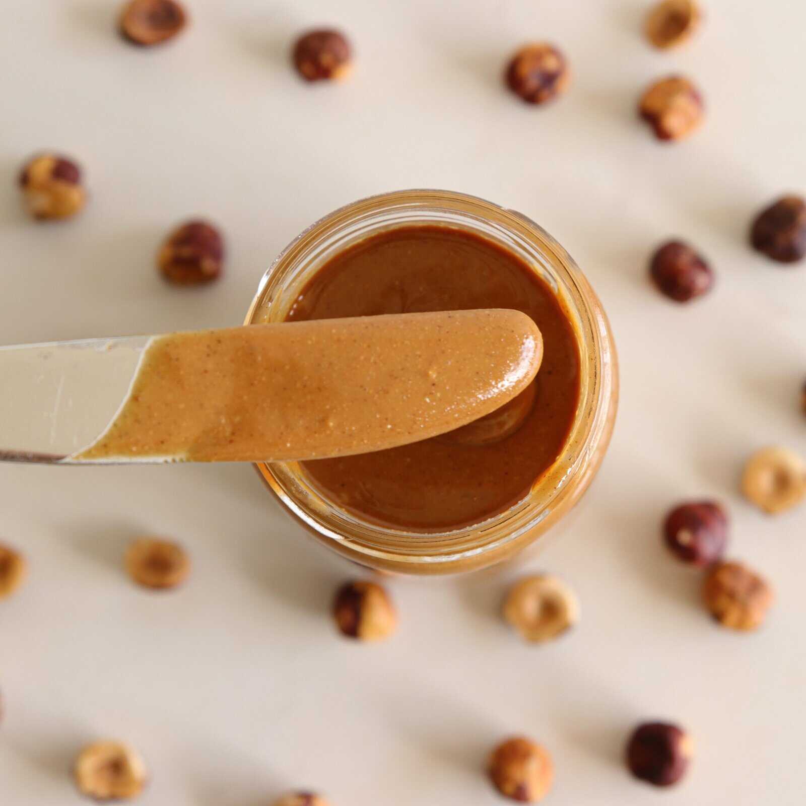 A knife with hazelnut spread rests over an open jar, surrounded by scattered whole hazelnuts on a light background.