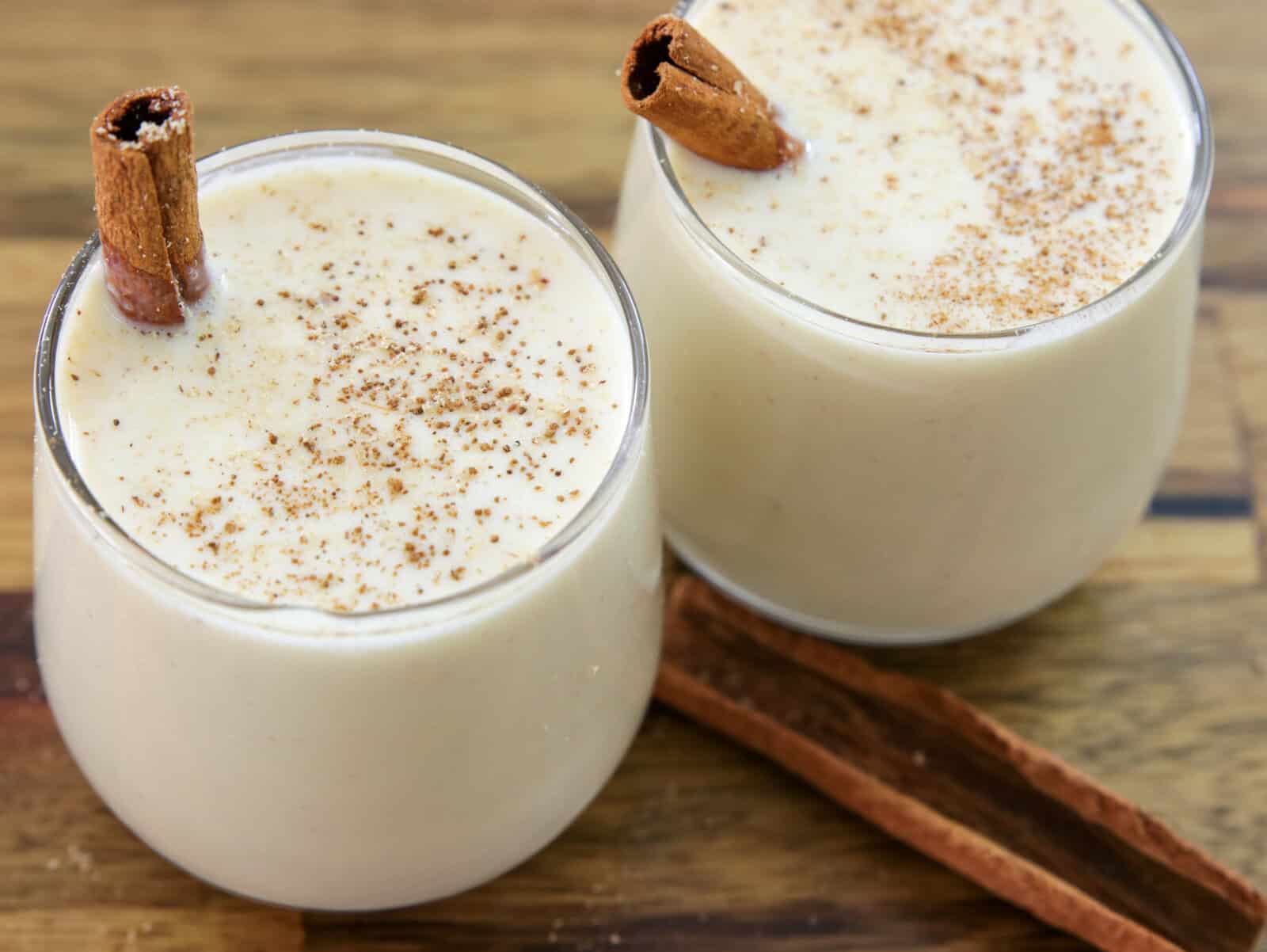 two short, rounded glass cups filled with a creamy, pale ivory drink that looks like classic eggnog. Each glass is garnished with a cinnamon stick resting on the rim, and the surface of the drink is lightly dusted with ground cinnamon or nutmeg. The glasses sit on a rustic wooden surface, with extra cinnamon sticks placed nearby, adding a warm, cozy, holiday feel. The eggnog appears thick, smooth, and rich, with a clean, simple presentation that highlights its creamy texture and festive spices.
