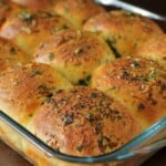 Golden brown garlic and herb dinner rolls, topped with chopped herbs and seasoning, arranged closely together in a glass baking dish.