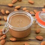 A glass jar of creamy almond butter with an orange-rimmed lid sits open on a wooden surface, surrounded by whole almonds scattered around it.