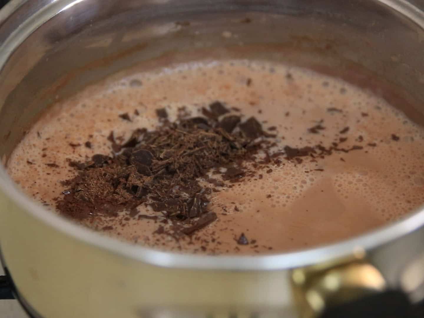 A saucepan filled with a creamy, light brown liquid topped with melting chunks and shavings of dark chocolate, likely hot chocolate being prepared on a stovetop.