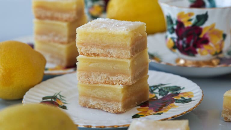 Three stacked lemon bars with powdered sugar on top sit on a floral plate, surrounded by whole lemons and a decorative teacup in the background. The lemon bars have distinct layers and a crumbly base.