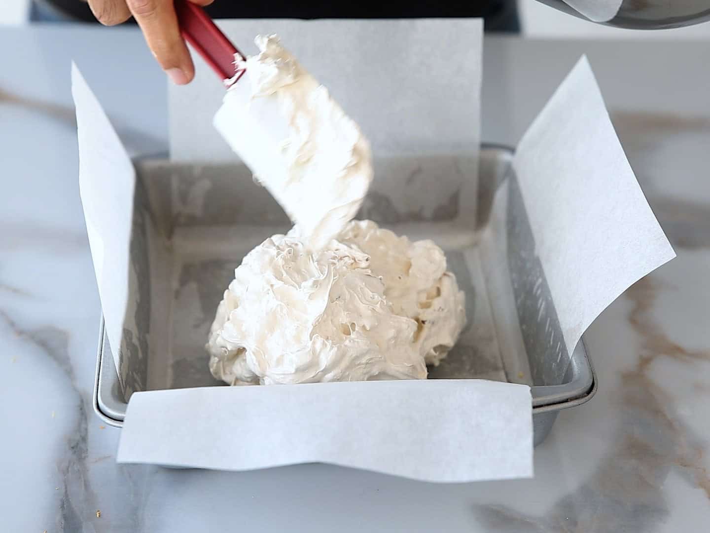 A hand uses a spatula to spread a thick, white mixture into a square baking pan lined with parchment paper on a marble surface.