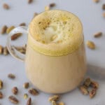 A glass mug filled with frothy pistachio chai latte, topped with crushed pistachios. Whole pistachio nuts are scattered on the light-colored surface around the mug.