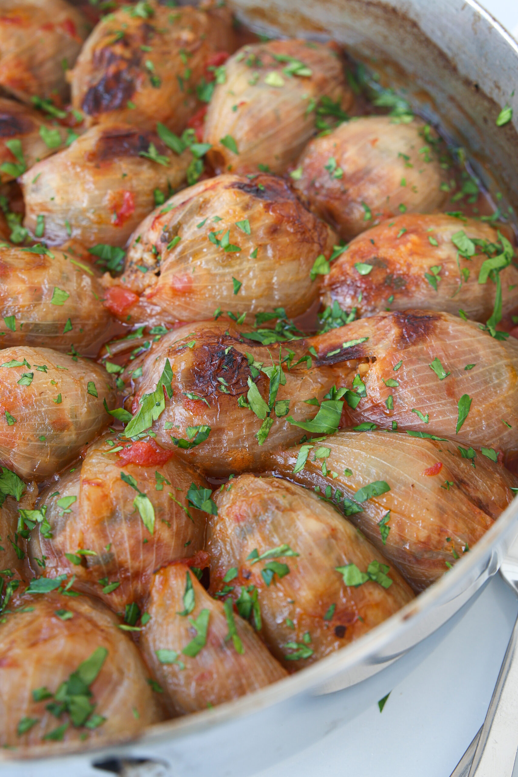 A close-up of stuffed onions baked in a pan, topped with chopped fresh herbs and lightly browned on top, with visible pieces of tomato in the sauce.