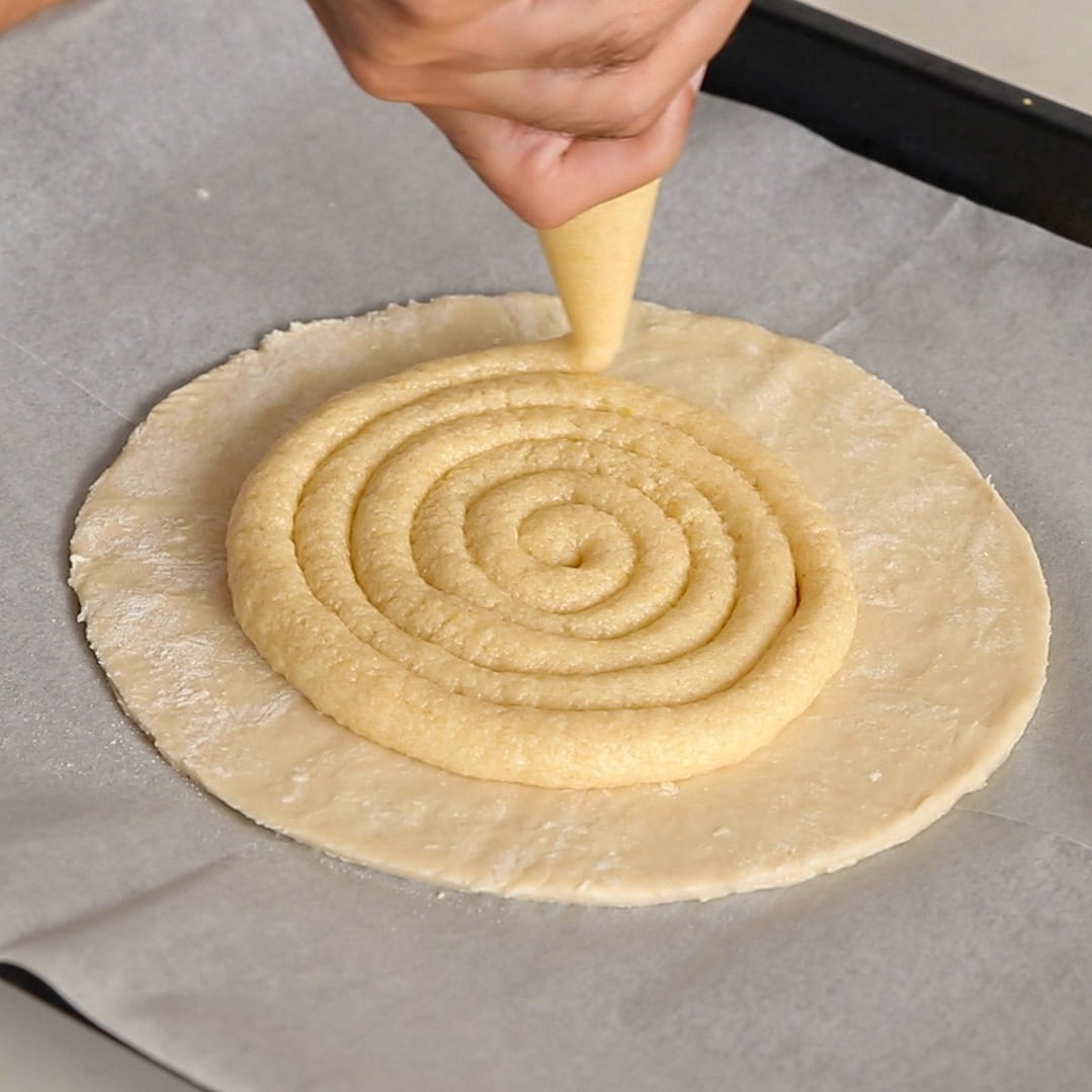 A hand uses a piping bag to pipe a spiral of filling onto a round sheet of dough placed on parchment paper.