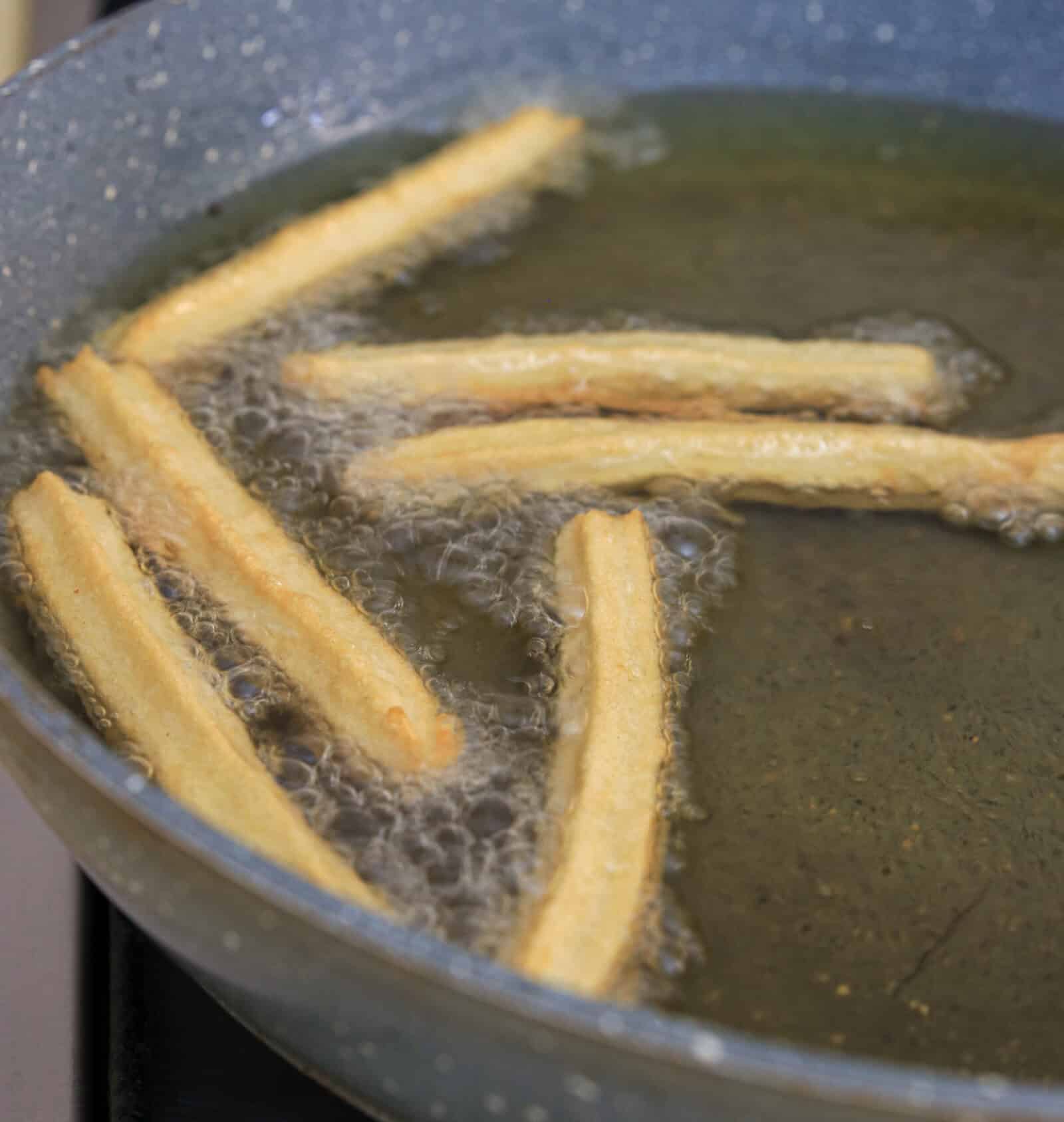 Churros frying in hot oil inside a large pan, with bubbles forming around the dough as they cook.