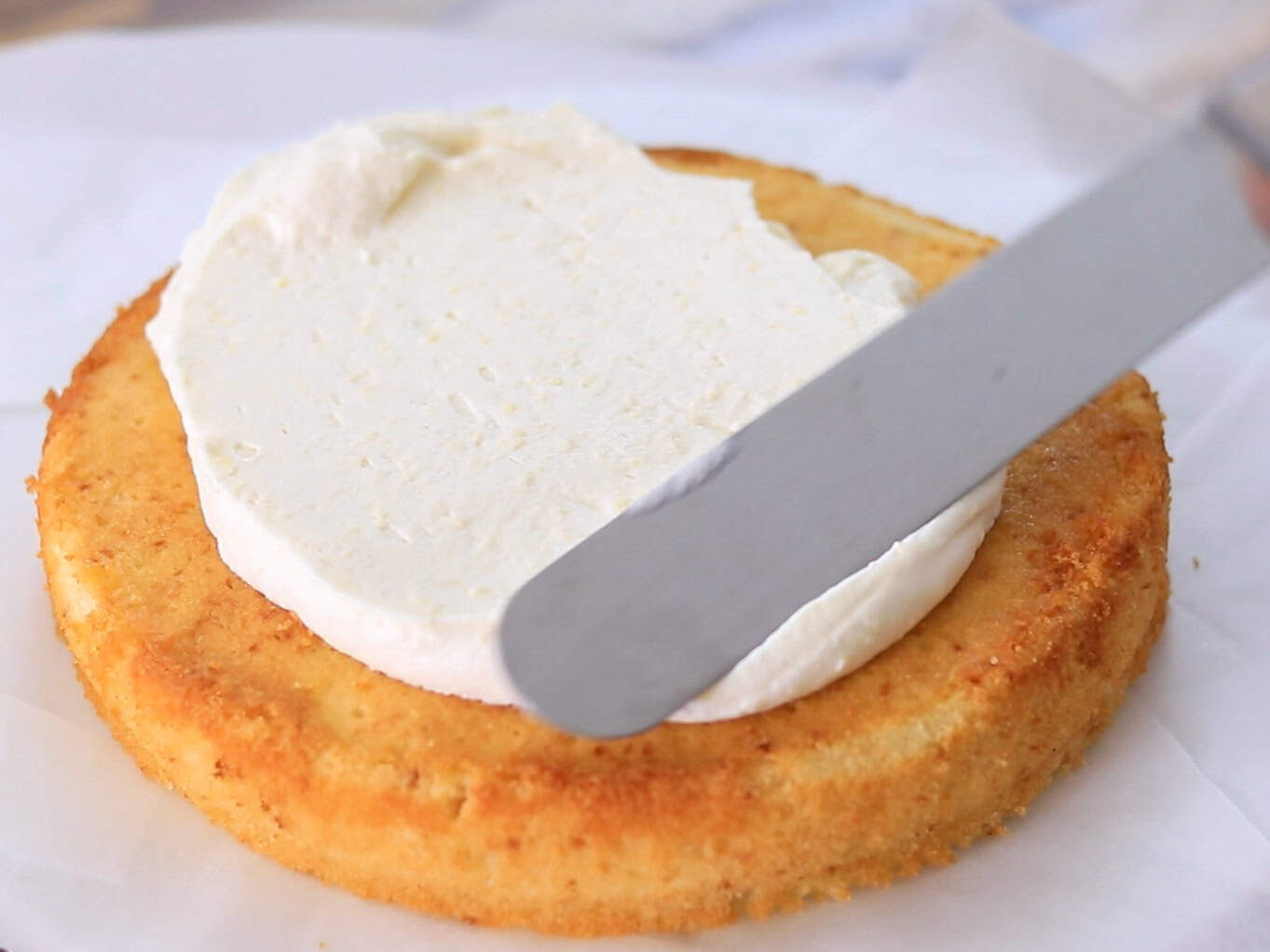 A close-up of a cake layer being spread with white frosting using an offset spatula on a piece of parchment paper.