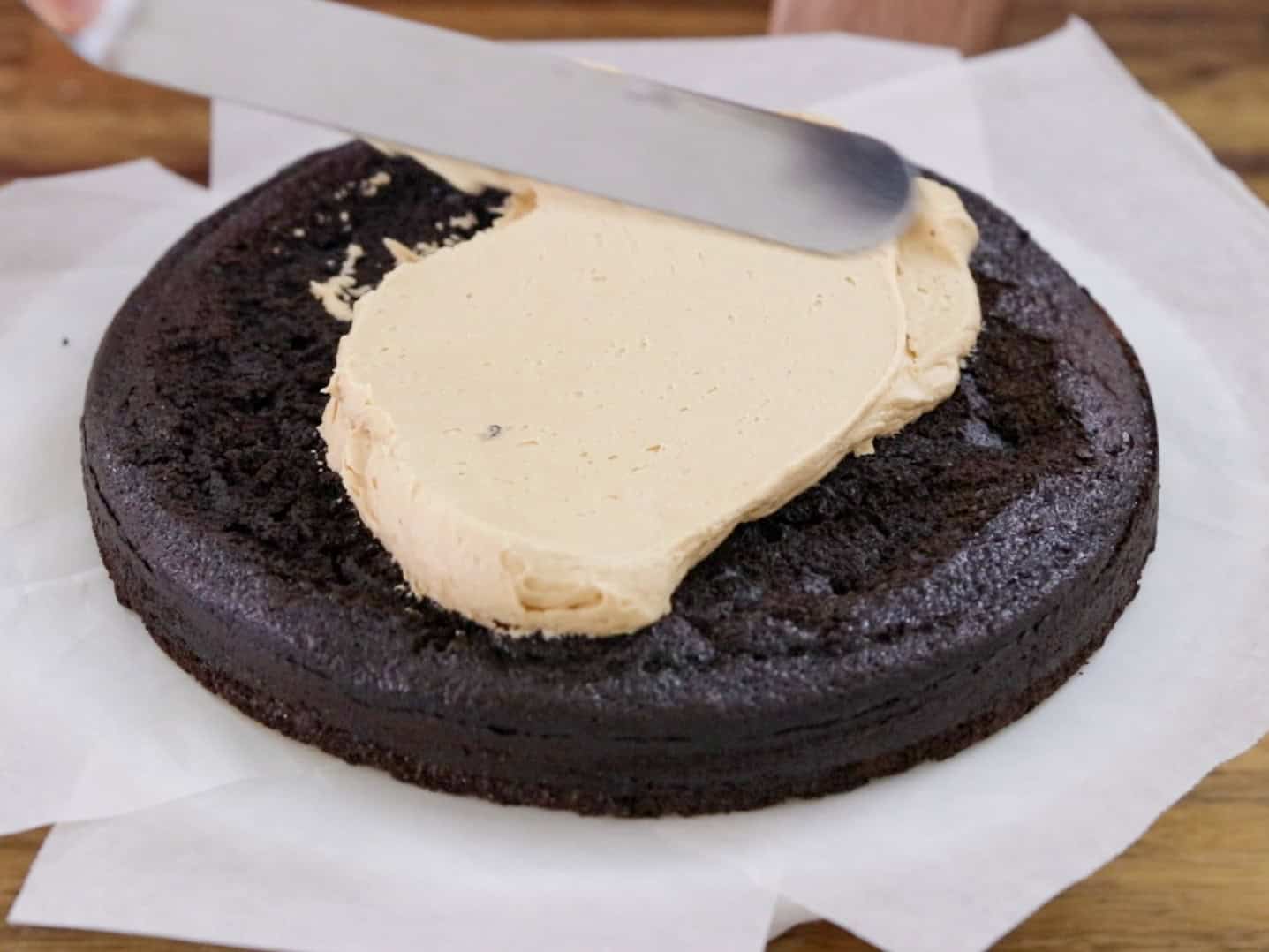 A metal spatula spreads light brown frosting over a round, unfrosted chocolate cake layer resting on parchment paper on a wooden surface.