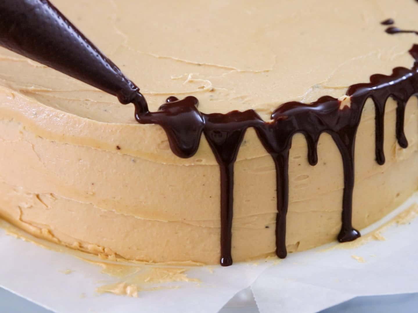 A close-up of a cake with light brown frosting, as chocolate ganache is piped along the edge, creating a drip effect down the sides.