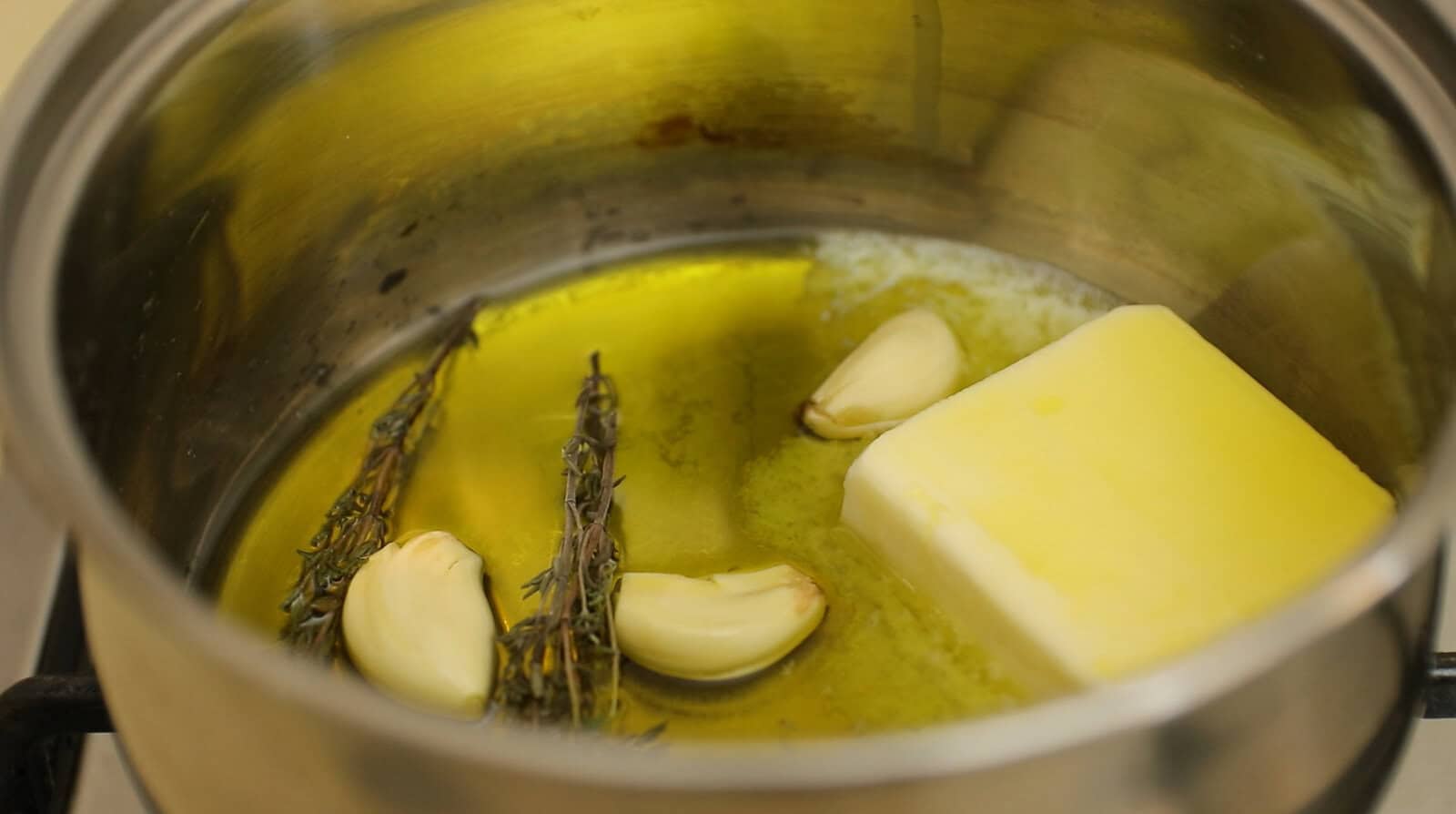 A saucepan with olive oil, a stick of butter, garlic cloves, and fresh thyme sprigs being heated together on a stove.