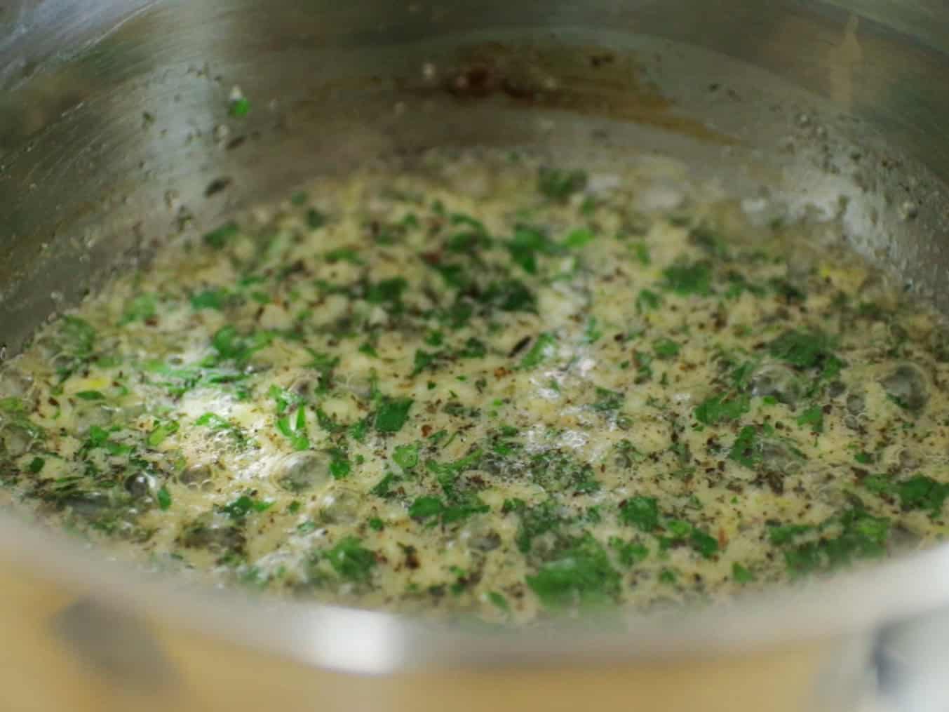 A close-up of a metal pot with a creamy mixture simmering, filled with chopped green herbs and seasoning, creating a textured and bubbling surface.