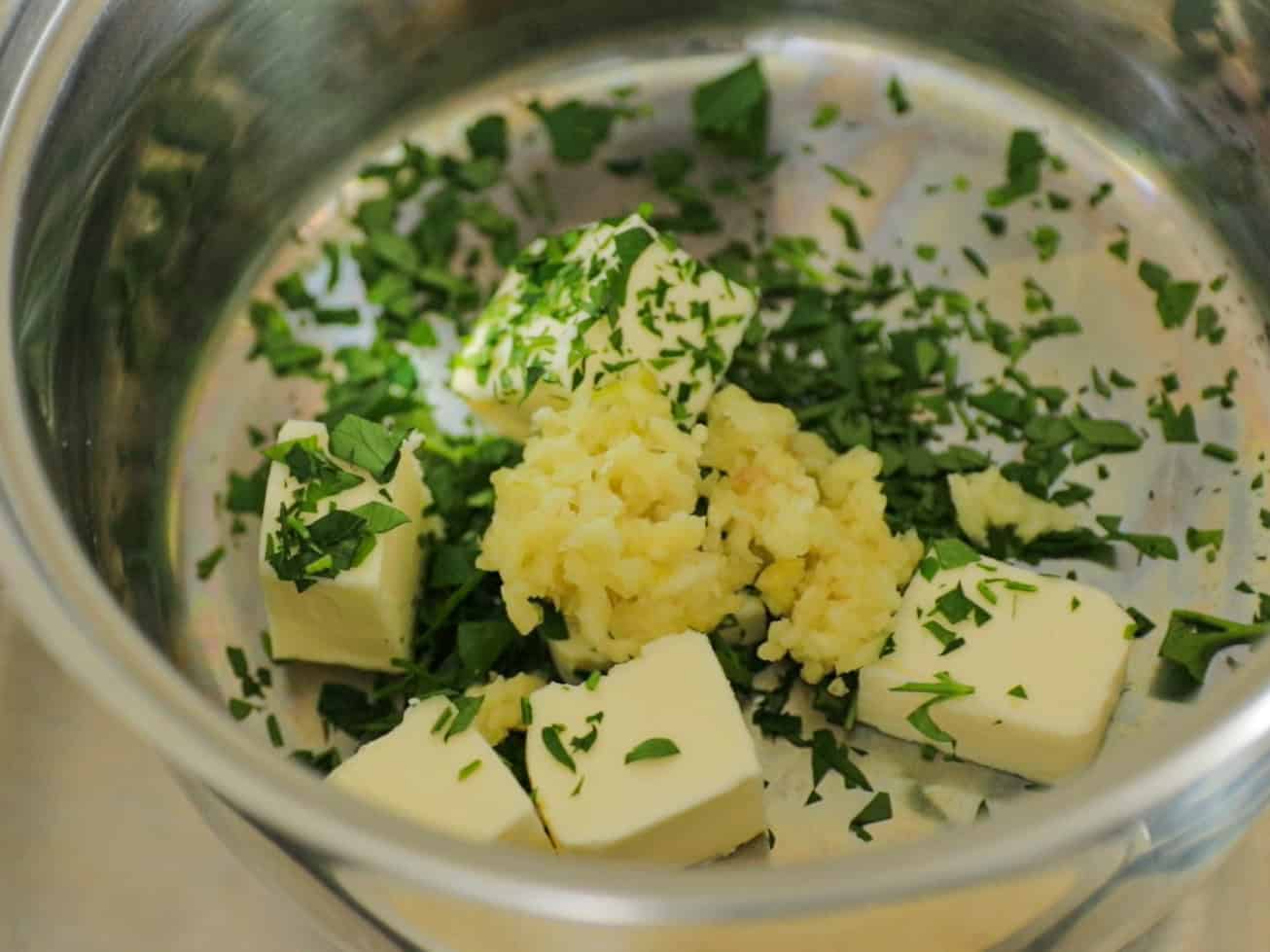Cubes of butter, chopped fresh parsley, and minced garlic are placed in a metal mixing bowl, ready to be combined.