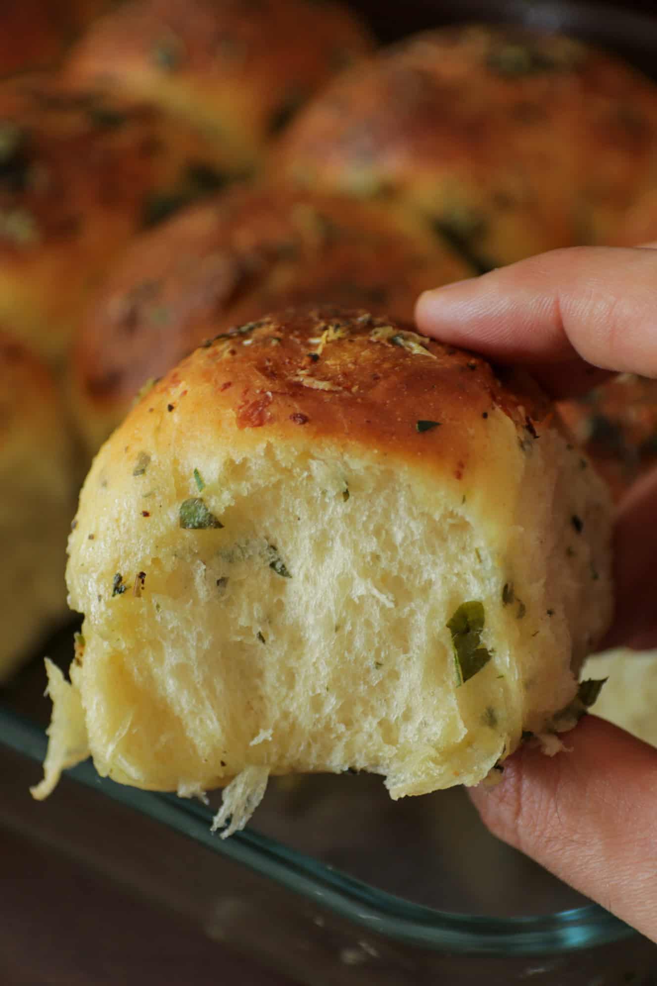 A hand holding a soft, golden-brown garlic bread roll topped with herbs, with more bread rolls visible in the background.