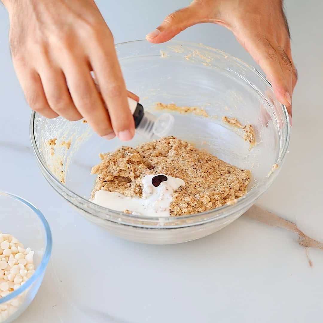 A person adds gel food coloring to a bowl of cookie dough mixture on a light countertop, with a bowl of white chocolate chips nearby.
