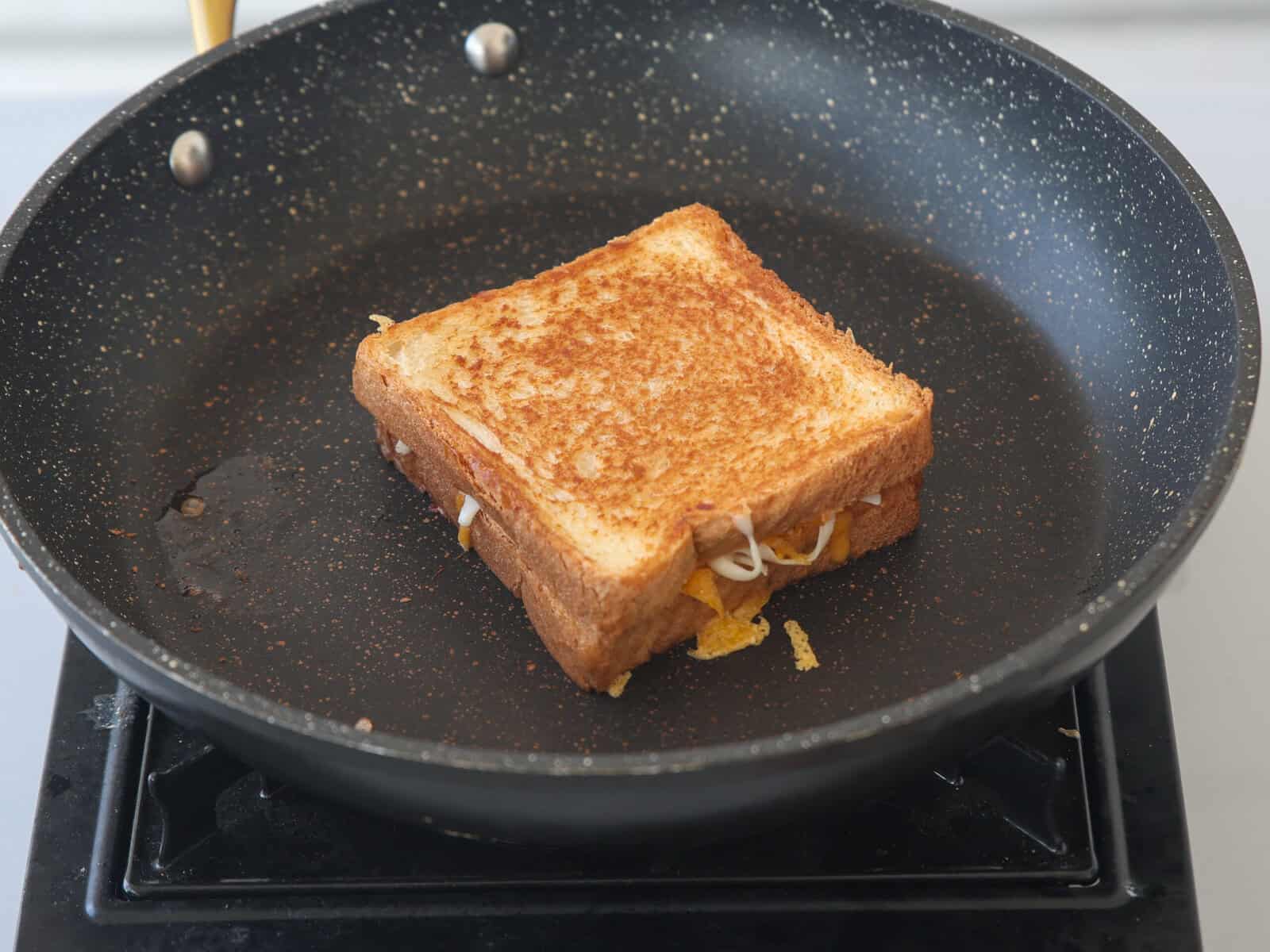 A grilled cheese sandwich with melted cheese is cooking in a nonstick frying pan on a stovetop.