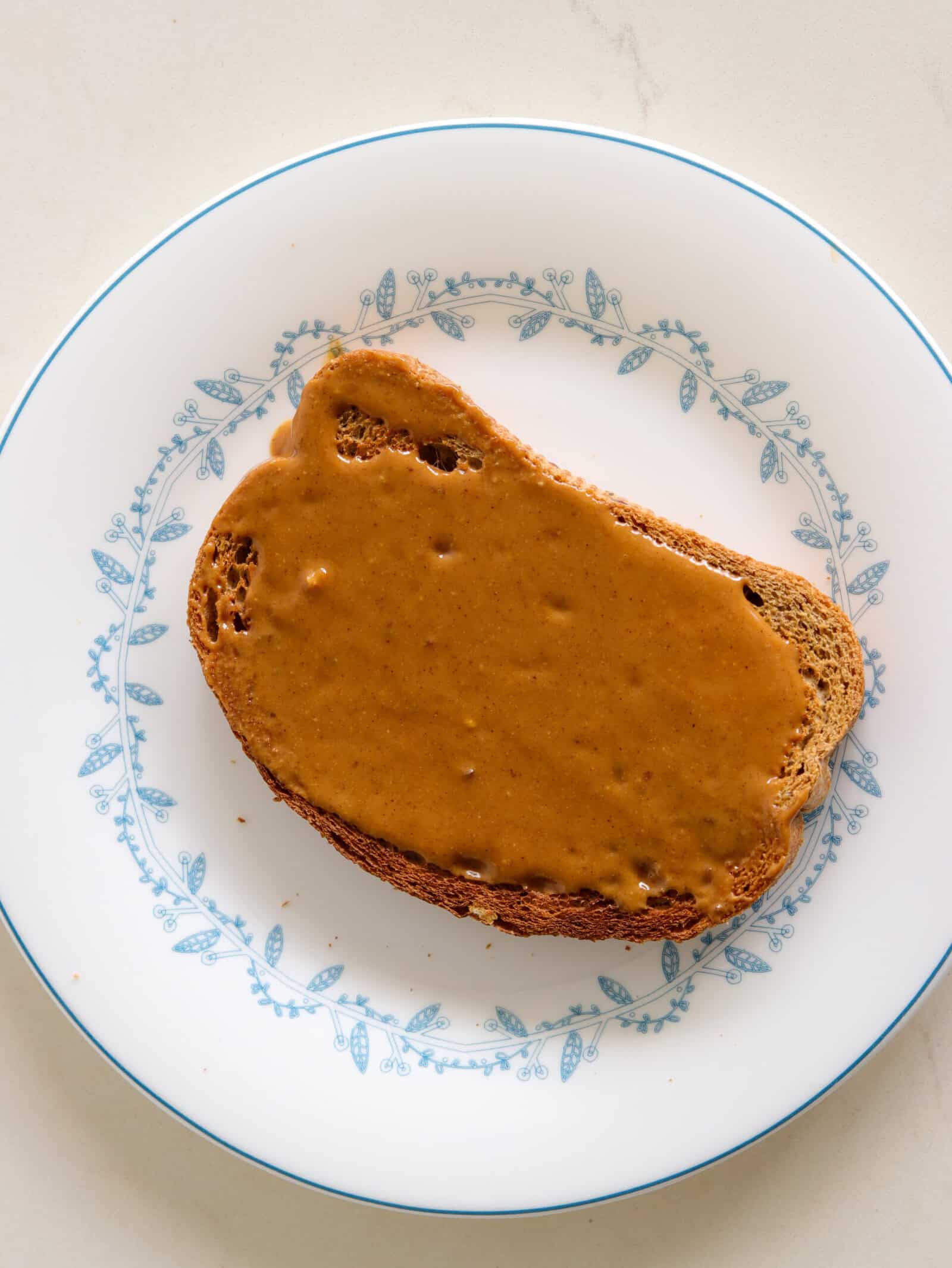 A slice of brown bread with a layer of hazelnut butter on top, placed on a white plate with a blue floral pattern, shown from above.
