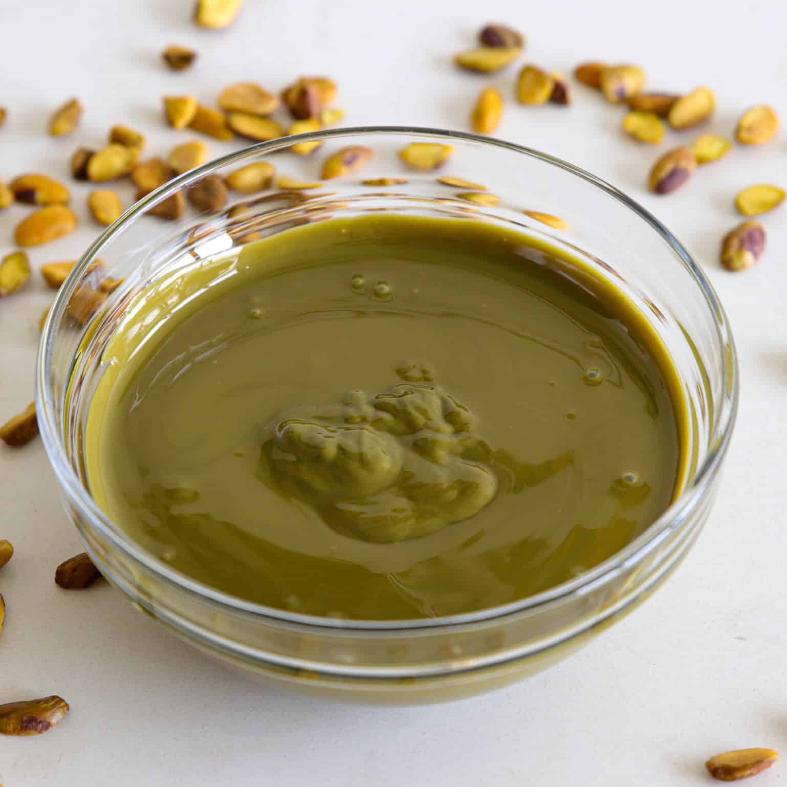 A clear glass bowl filled with smooth green pistachio butter sits on a white surface, surrounded by scattered whole pistachio nuts.
