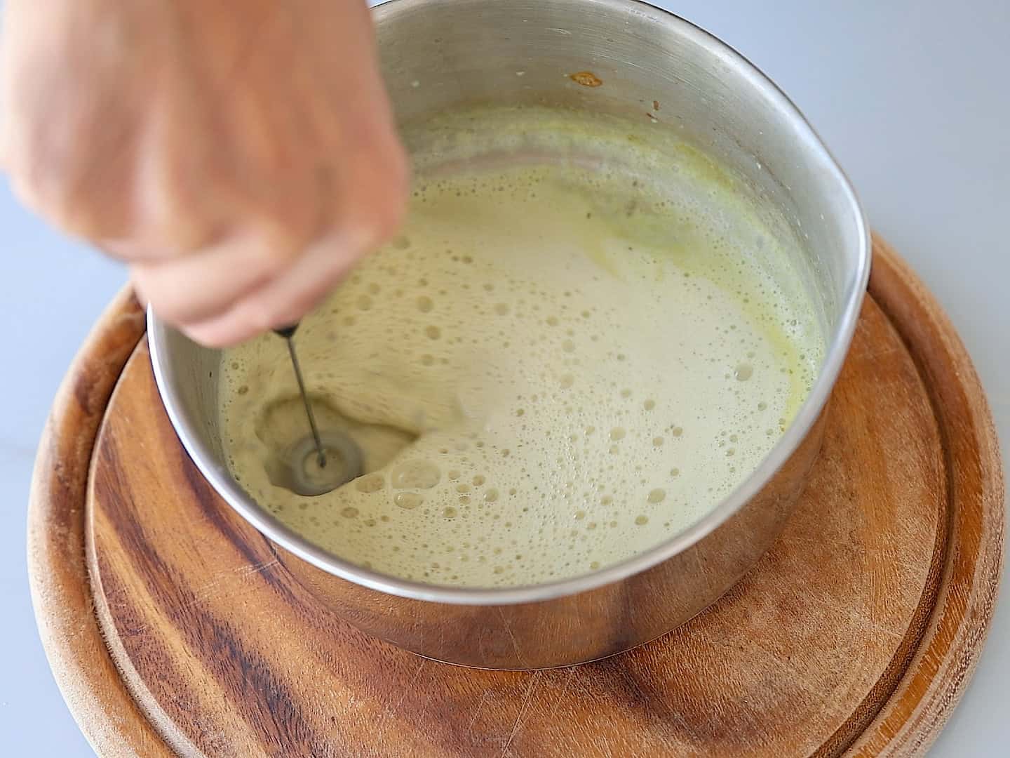 A hand uses a small electric whisk to froth a light green liquid in a metal bowl placed on a round wooden board.
