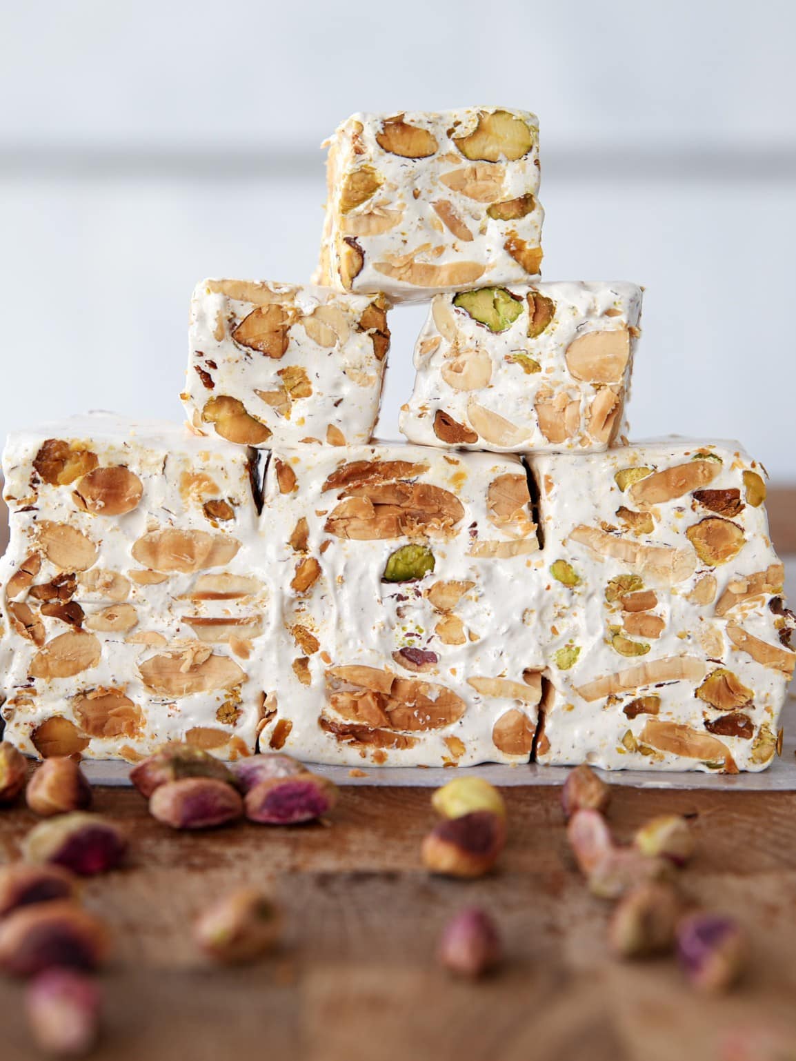 A stack of nut nougat (torrone) pieces, filled with pistachios and almonds, arranged on a wooden surface with whole pistachios scattered around.