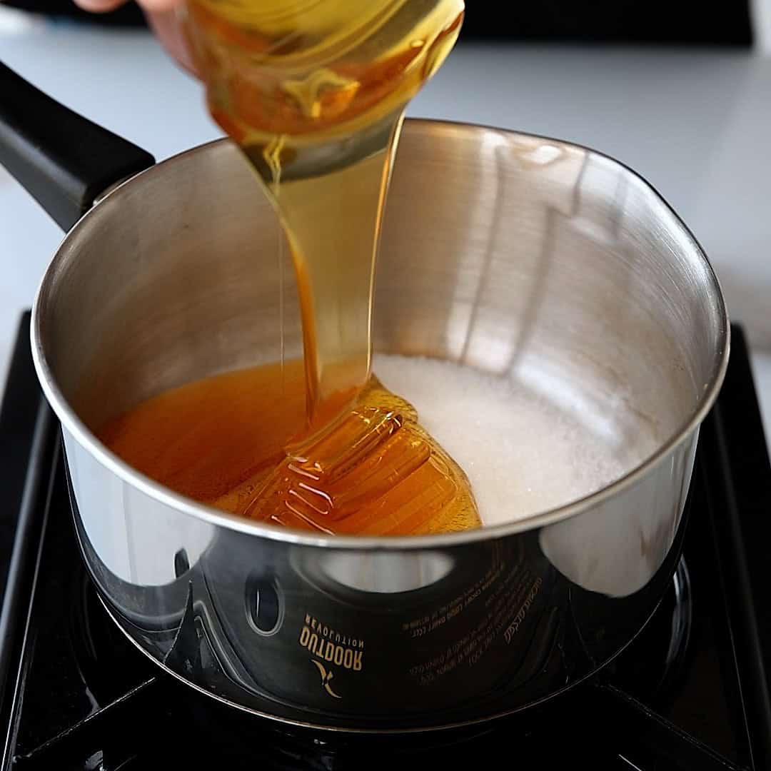 Honey is being poured from a jar into a saucepan containing white sugar, both starting to melt on a stovetop.