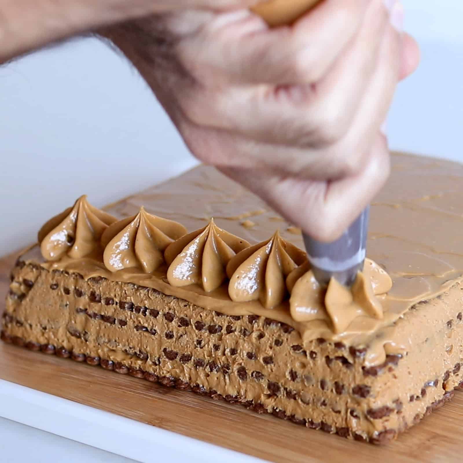 A hand uses a piping bag to decorate the edge of a rectangular cake with swirls of caramel-colored frosting. The cake rests on a wooden board and has a layered, chocolate-speckled appearance.