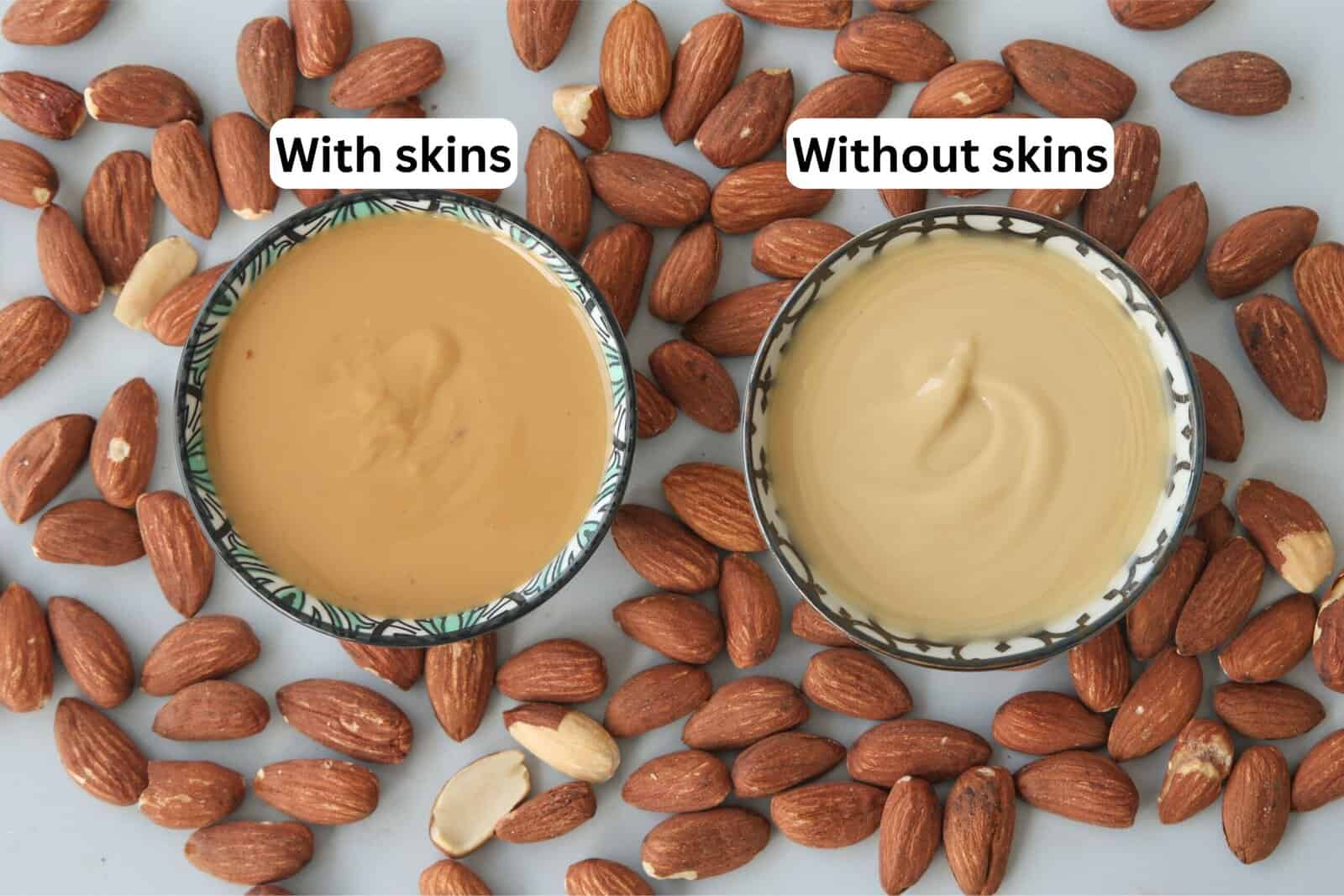 Two bowls of almond butter, one labeled “With skins” showing a darker spread, and one labeled “Without skins” showing a lighter spread, surrounded by whole almonds and a few almond pieces.
