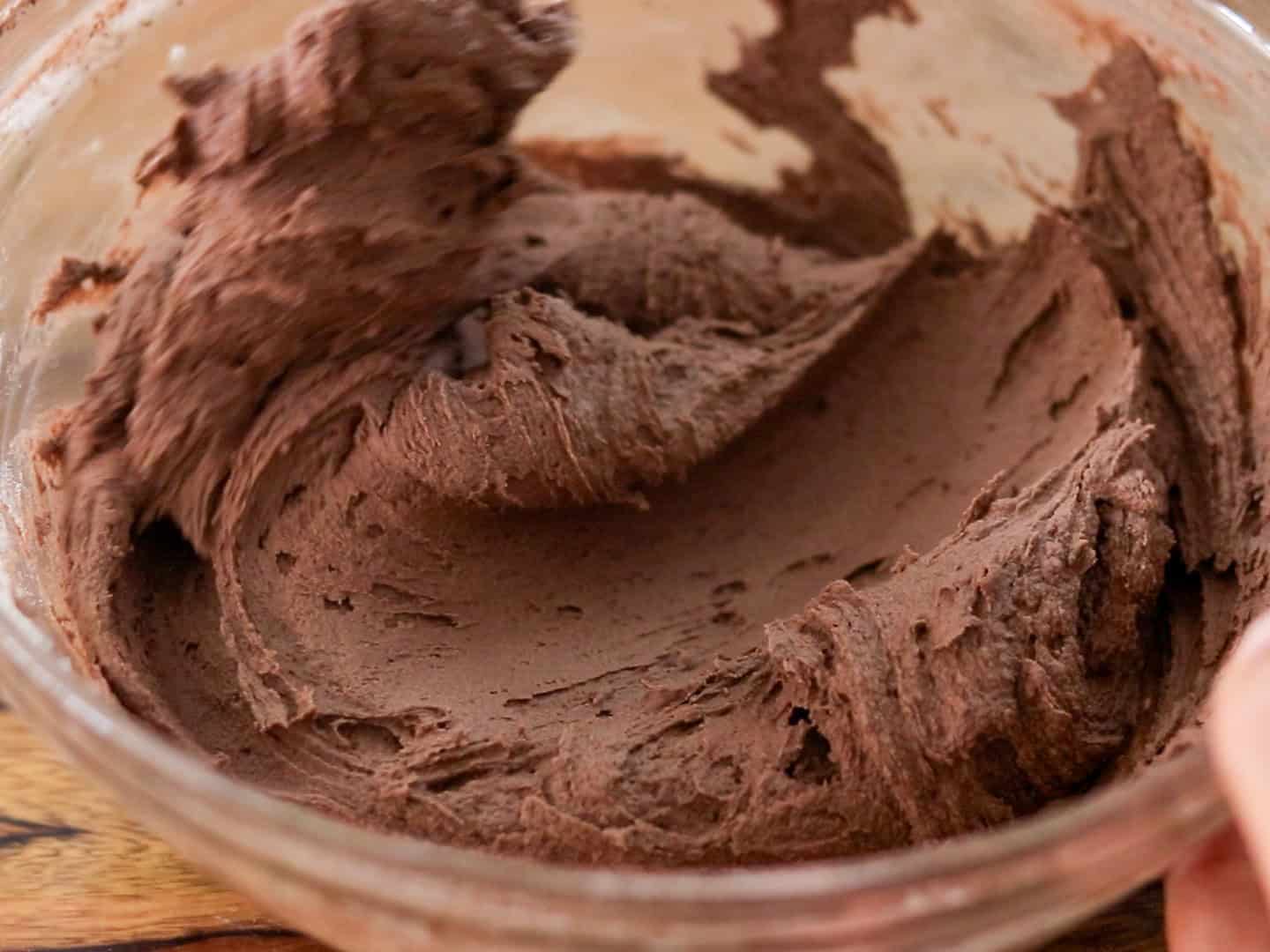 A glass bowl filled with thick, creamy chocolate batter being mixed, showing smooth, swirled texture on the sides of the bowl.