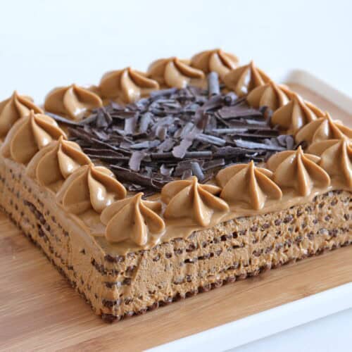 A square coffee-flavored cake with piped frosting swirls on top, decorated with chocolate shavings in the center, served on a wooden tray.