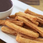 A plate of churros dusted with sugar sits next to a small bowl of chocolate dipping sauce on a white rectangular dish.